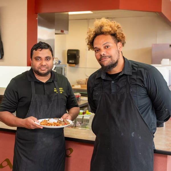Two male chefs in black aprons, one holding a plate of food, standing in a restaurant kitchen.