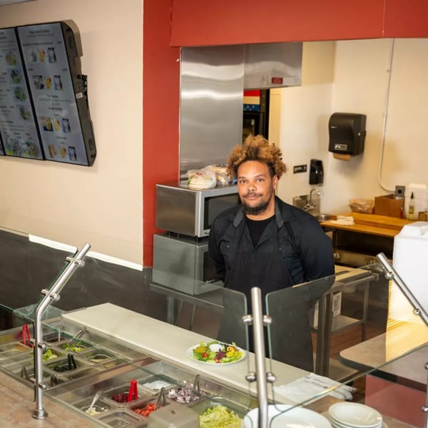 A chef stands behind a salad bar with a prepared salad and menu screens in the background.