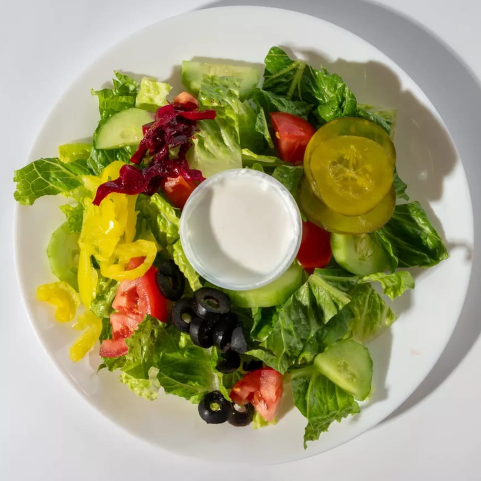 A fresh garden salad with lettuce, tomatoes, cucumbers, olives, peppers, and a side of dressing.