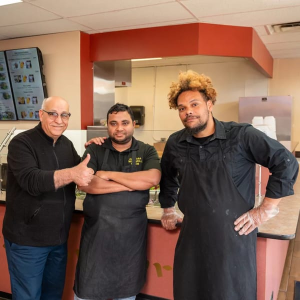 Three men, two in black aprons, stand smiling behind a counter, one giving a thumbs-up.